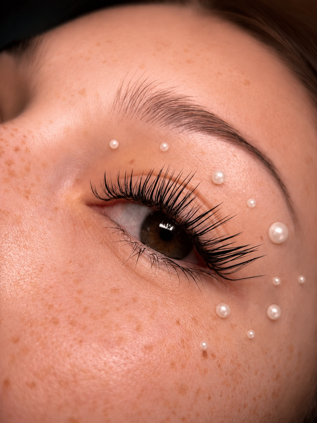 Detal stylizacji rzęs. Volume lashes plus perłowe aplikacje na skórze wokół oka, naturalne piegi modelki.